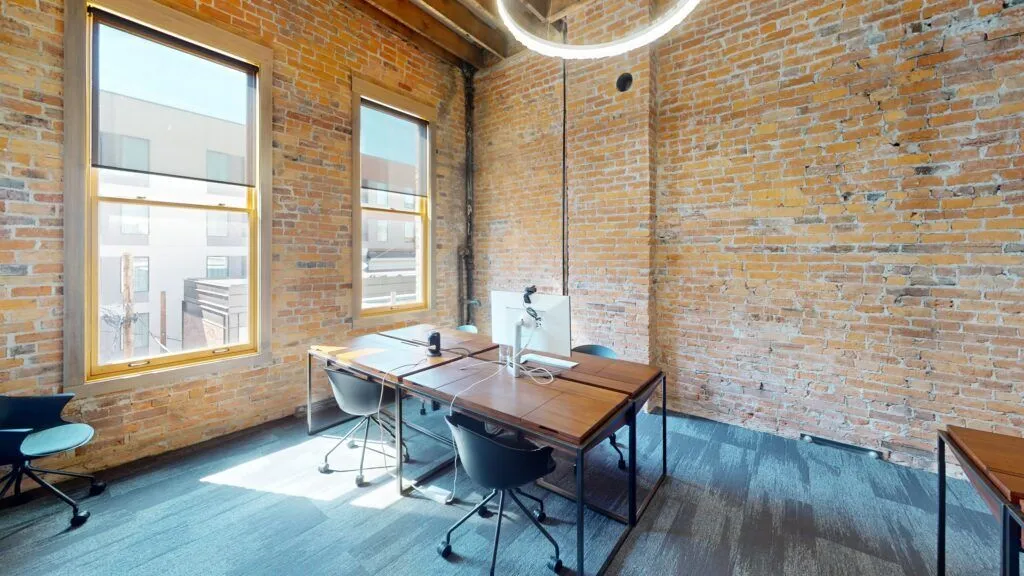 A modern office with exposed brick walls, large windows, four desks with chairs, a circular hanging light, and office supplies on the desks. Natural light fills the room—perfect for a Missoula Commercial Broker or any Missoula commercial real estate professional.