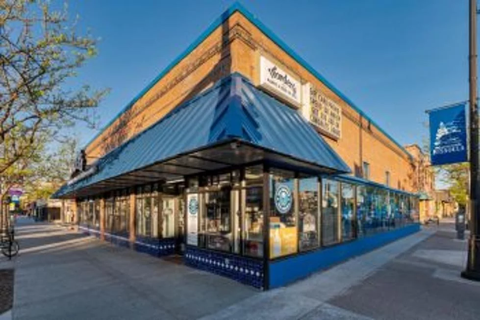 A corner view of a brick building with large glass windows and a blue awning, located on a city sidewalk under a clear blue sky—a perfect example for any Missoula Commercial Broker seeking inspiration. Signs include one that reads Midtown Global Market.