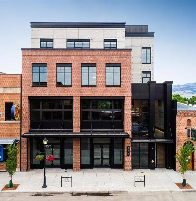 A modern, three-story brick building with large windows and black trim stands on a clean city sidewalk, surrounded by older structures and distant mountains—an ideal property for any Missoula Commercial Real Estate opportunity.