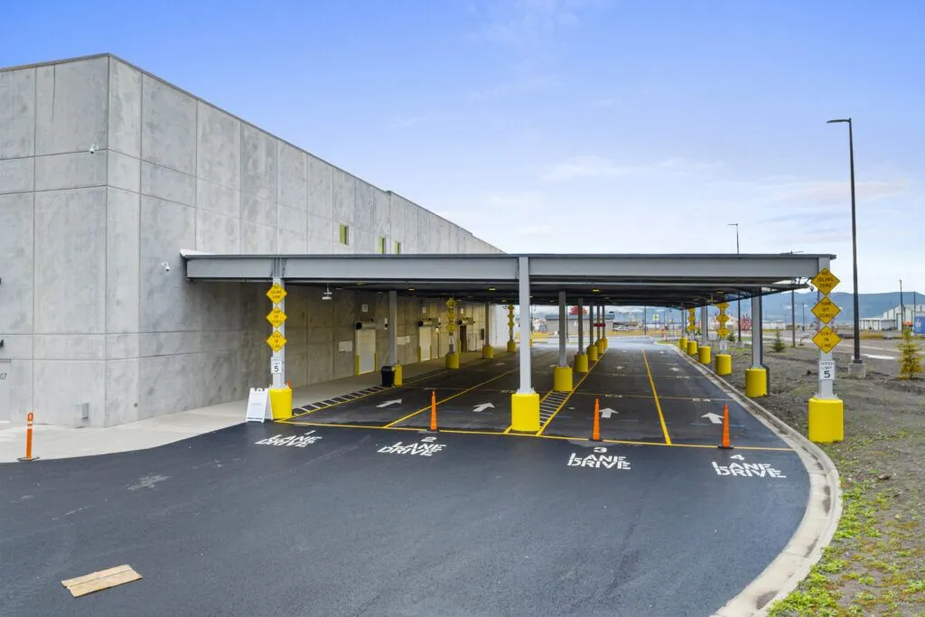 A covered drive-thru area at a modern, industrial-style building with multiple lanes marked DRIVE SLOW, yellow posts, and designated stops—perfectly suited for your needs by a trusted Missoula Commercial Property Broker—on a freshly paved surface under a blue sky.