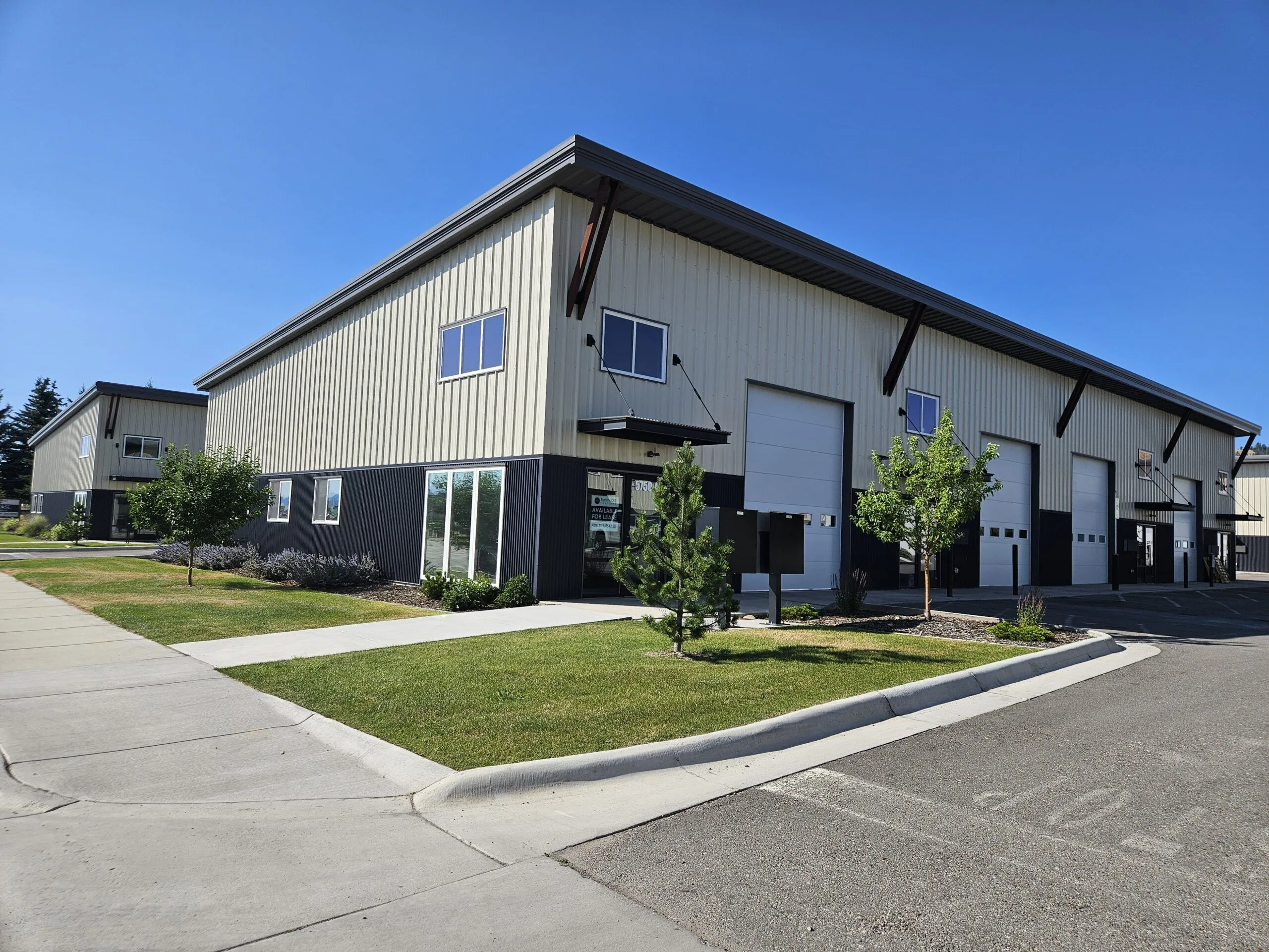 A modern industrial building with beige and black siding, large garage doors, and office windows—an ideal opportunity for a Missoula Commercial Broker—surrounded by grass, small trees, and a sidewalk under a clear blue sky.
