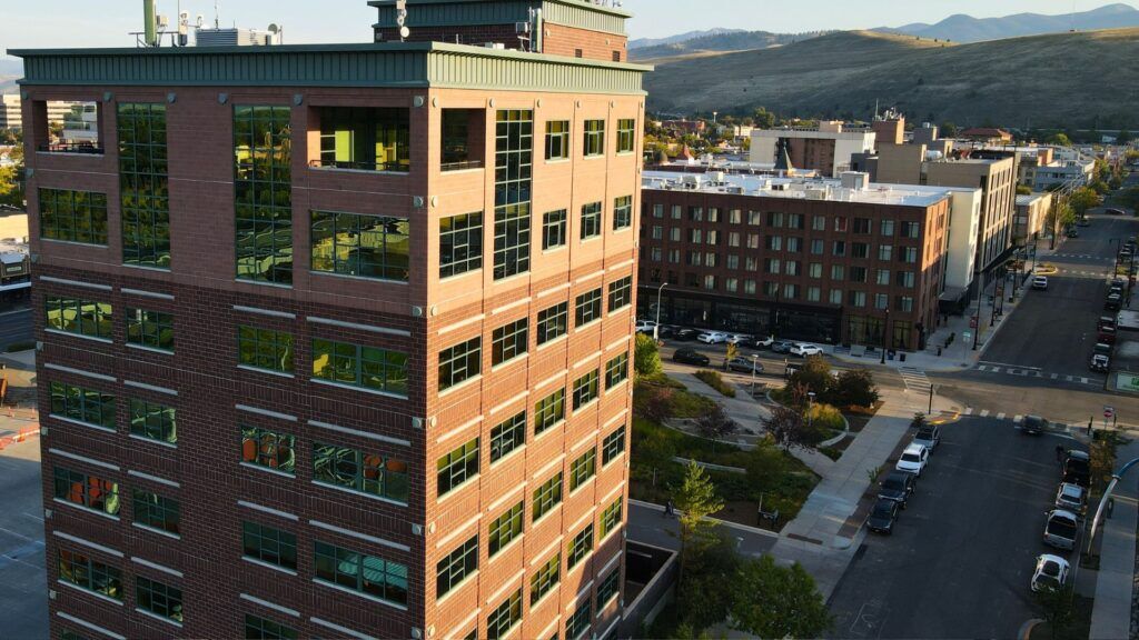 A tall brick office building with large windows stands at a street corner in a small city, perfect for a Missoula Commercial Real Estate venture. Other buildings, parked cars, and rolling hills are visible in the daylight background.