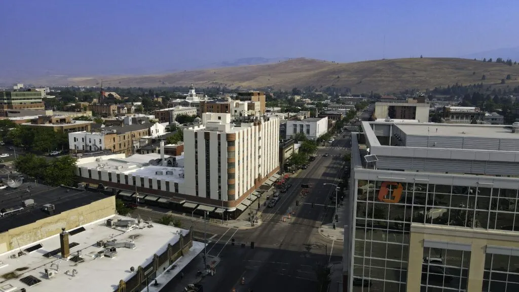 Aerial view of a mid-sized city with multi-story buildings, streets, and cars, surrounded by hills and trees under a hazy blue sky—perfect for those seeking Missoula Commercial Real Estate opportunities.