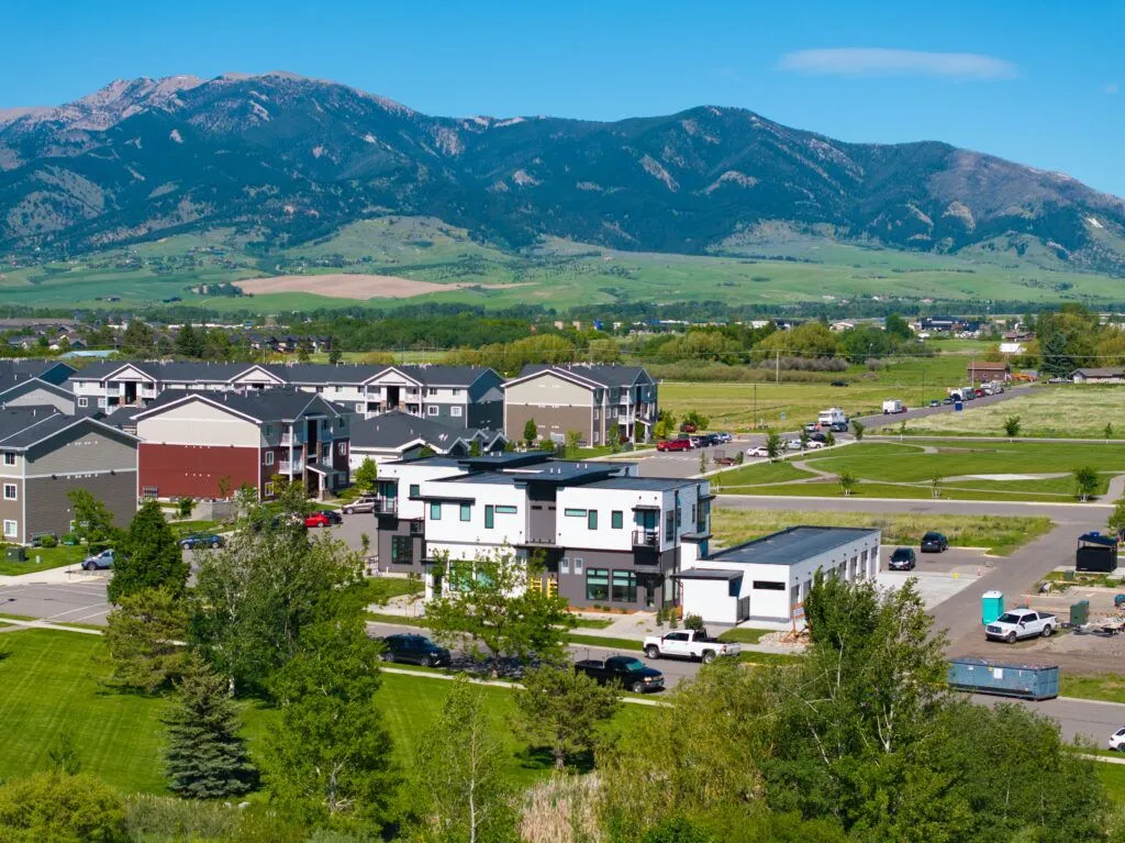 A small suburban neighborhood with modern homes and green lawns sits in front of tree-covered mountains under a clear blue sky—an inviting area explored by any Missoula Commercial Property Broker seeking prime locations.