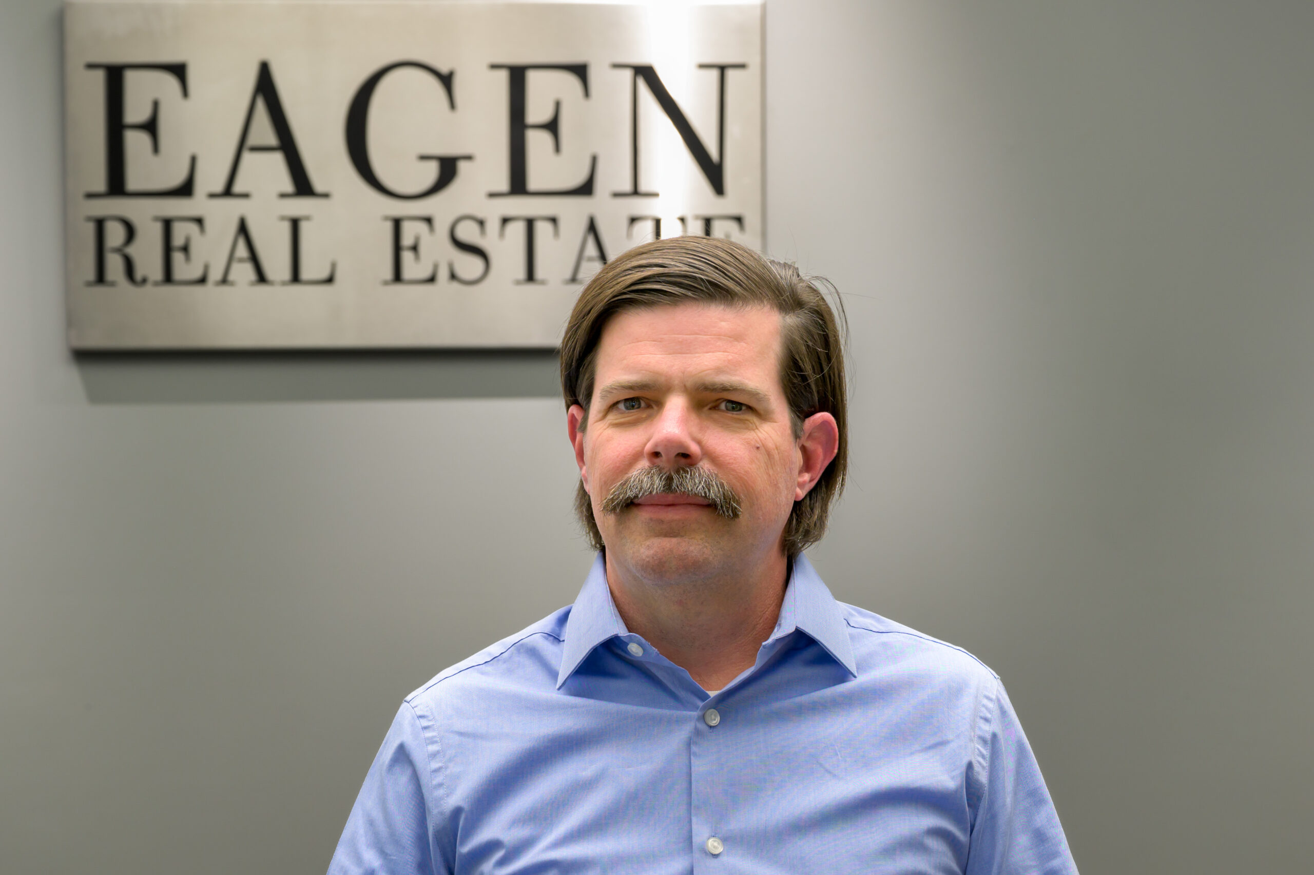 A man with a mustache and neatly combed hair stands in front of a sign that reads Eagen Real Estate. Wearing a light blue button-up shirt, he faces the camera, representing himself as your trusted Missoula Commercial Broker.