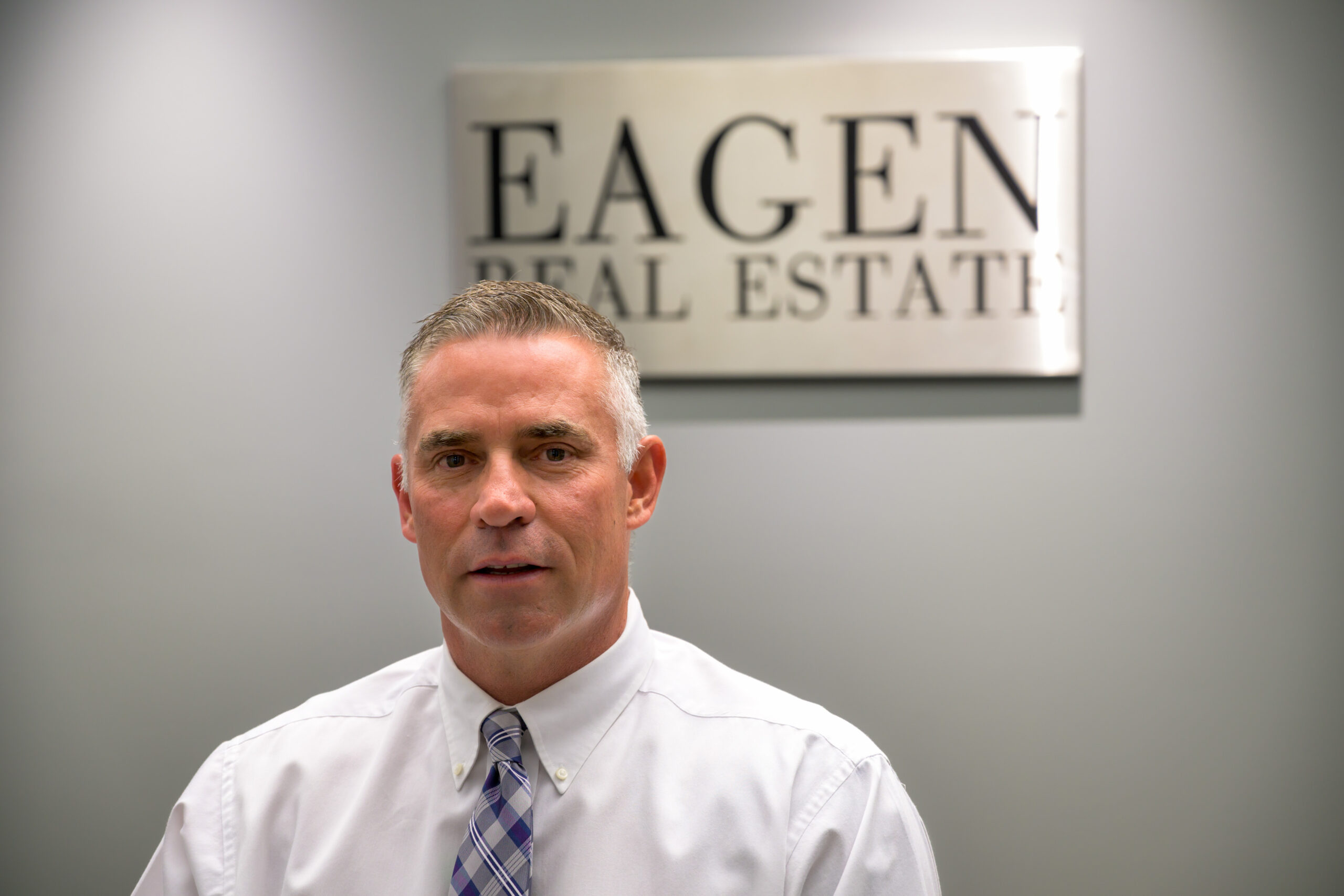 A man in a white dress shirt and plaid tie sits in front of a wall sign that reads EAGEN REAL ESTATE, presenting a neutral, professional setting—ideal for a Missoula Commercial Real Estate broker.